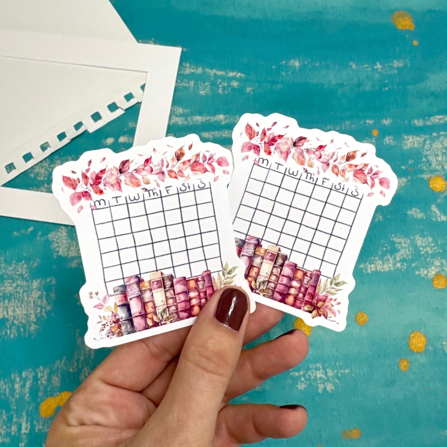 Hand holding two decorative planner stickers with floral and grid designs on a textured blue background.