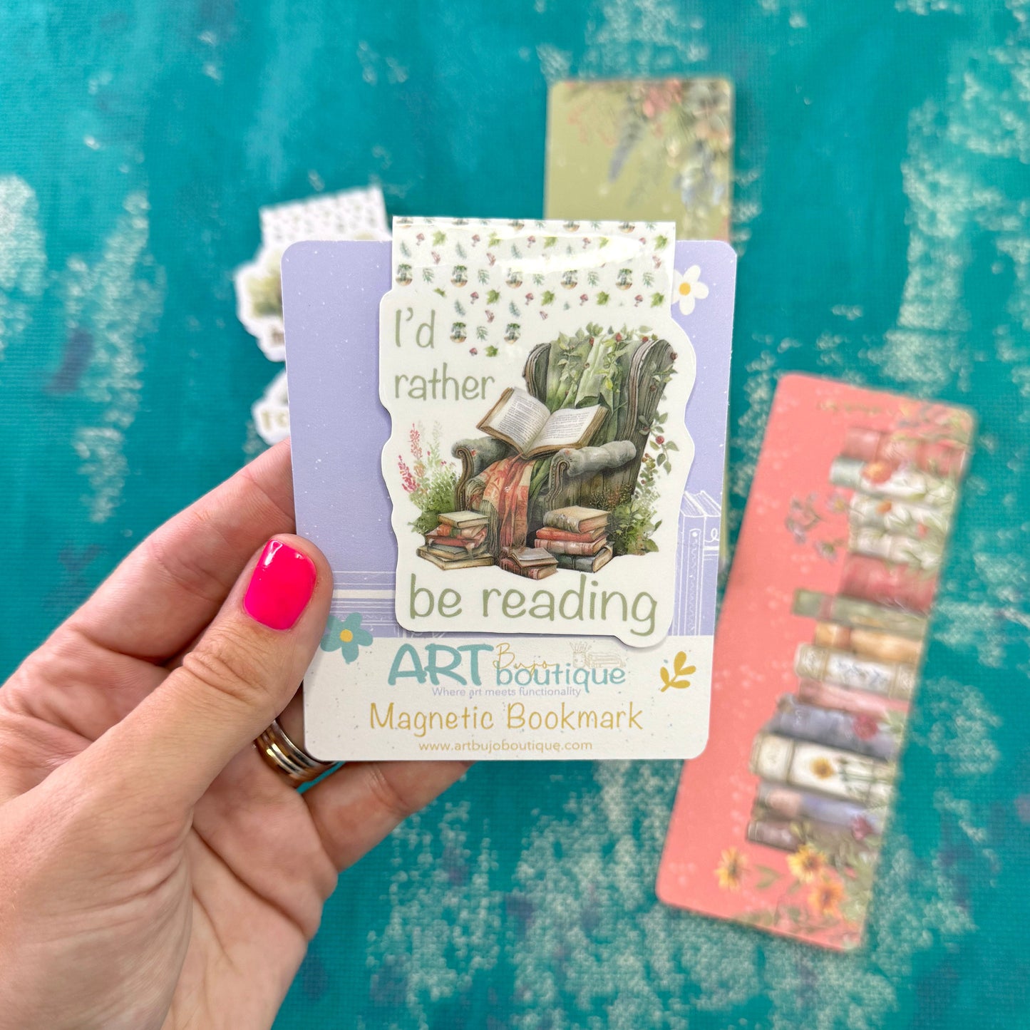 A hand with bright pink nail polish holds a lavender and white 'be reading' magnetic bookmark card featuring an illustration of a person reading in a chair. In the background, another pink card with bookmarks is visible against a textured teal backdrop.