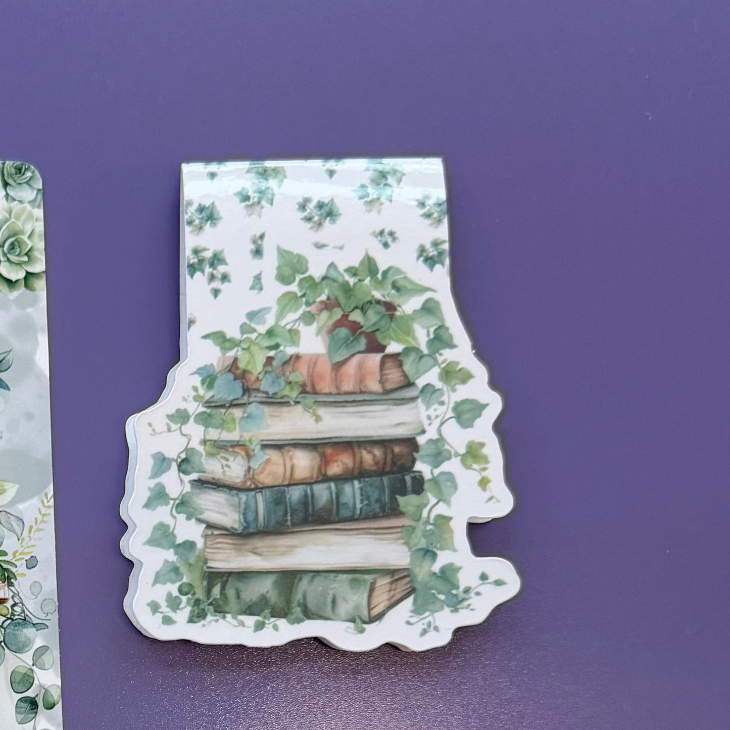 Four decorative bookmarks with watercolor illustrations of book stacks, vines, and a small forest scene, arranged on a purple surface with stationery props.