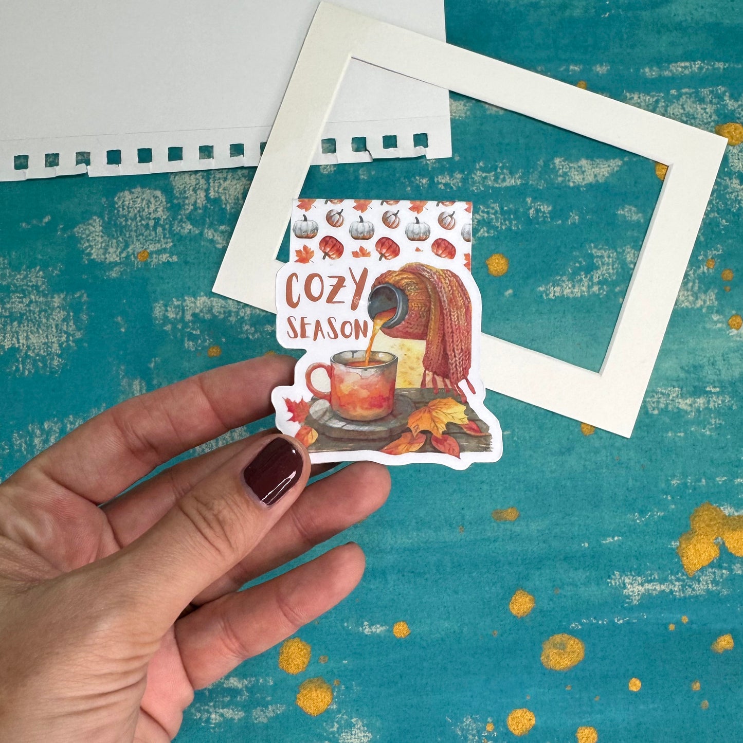 Hand holding a 'Cozy Season' sticker with a cup of coffee and autumn leaves on a textured blue background.