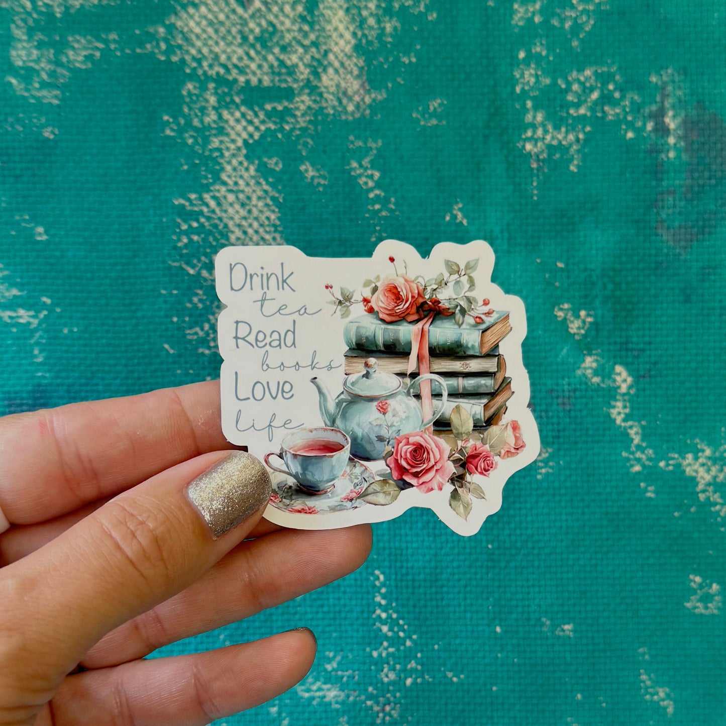 Hand holding a decorative sticker with tea and book-themed design against a textured teal background