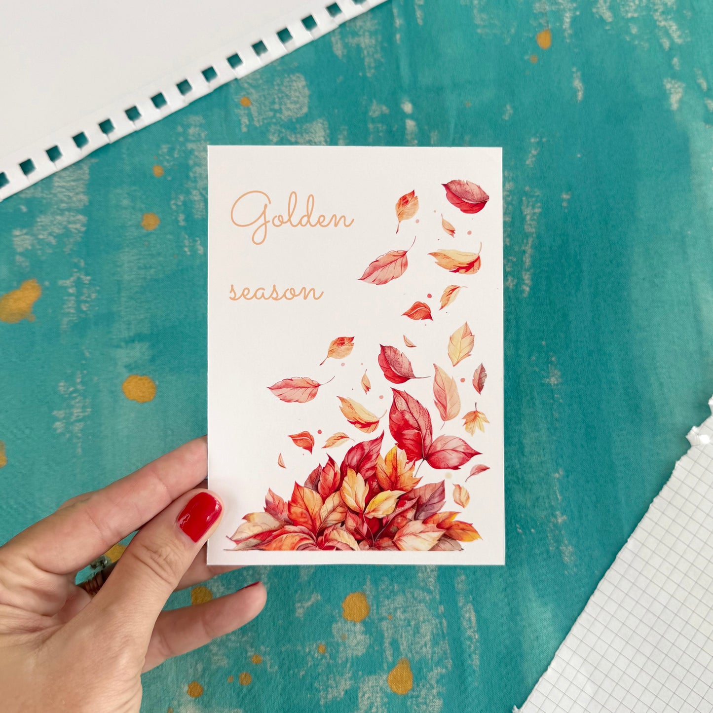 Card with autumn leaves and 'Golden season' text held by a hand on a textured teal background.