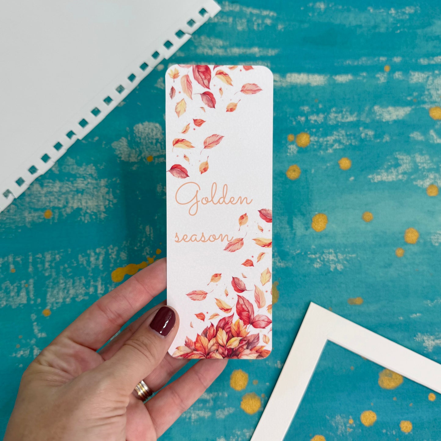 Hand holding a bookmark with autumn leaf design and 'Golden season' text on a blue textured background.