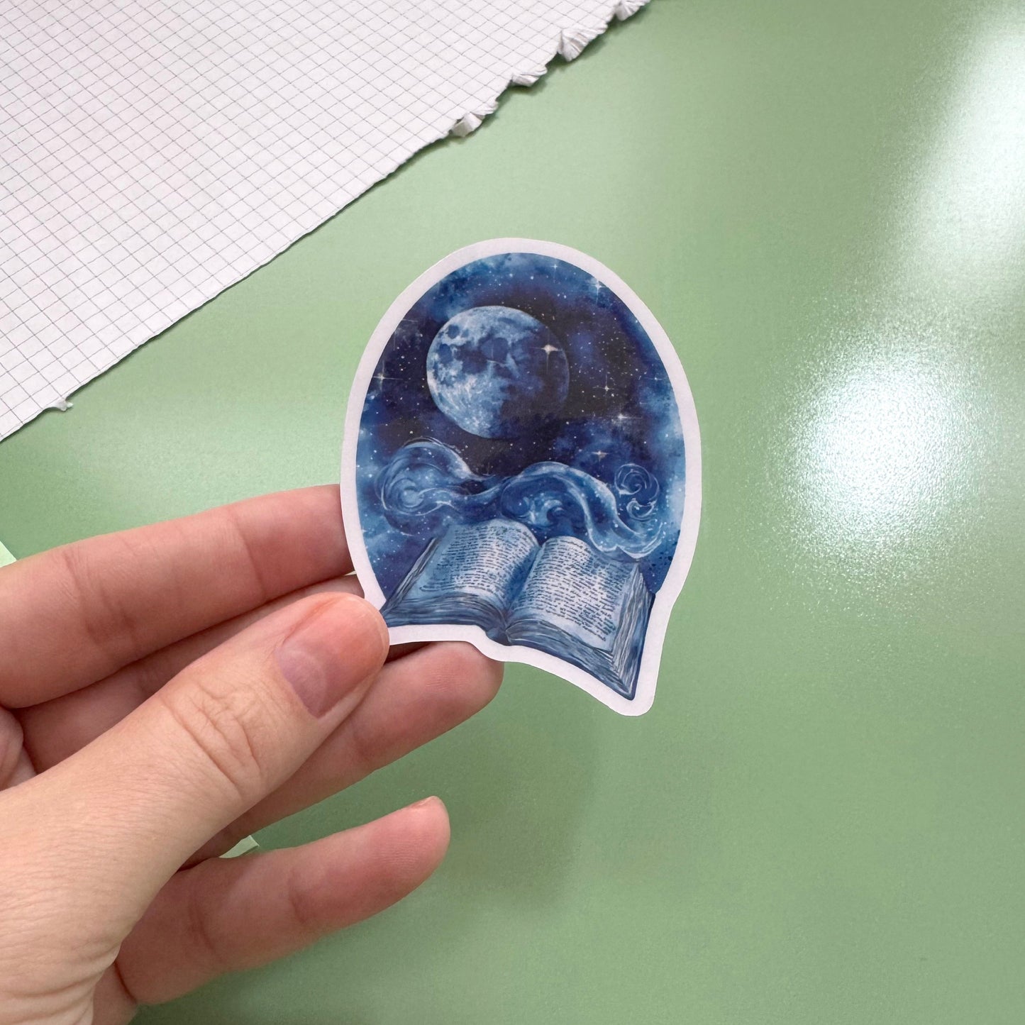 A hand holds a die-cut sticker featuring a blue night-sky illustration with a moon and open book, against a green tabletop background.