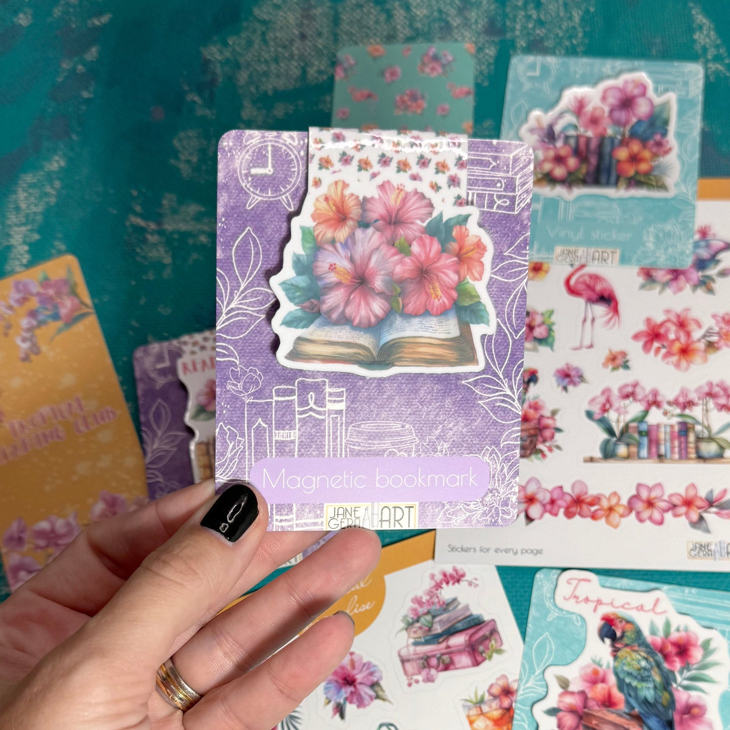 A hand with black nail polish holds several decorative sticker sheets featuring colorful vintage-style illustrations, including flowers, a parrot, a basket, and a book, over a teal patterned surface.