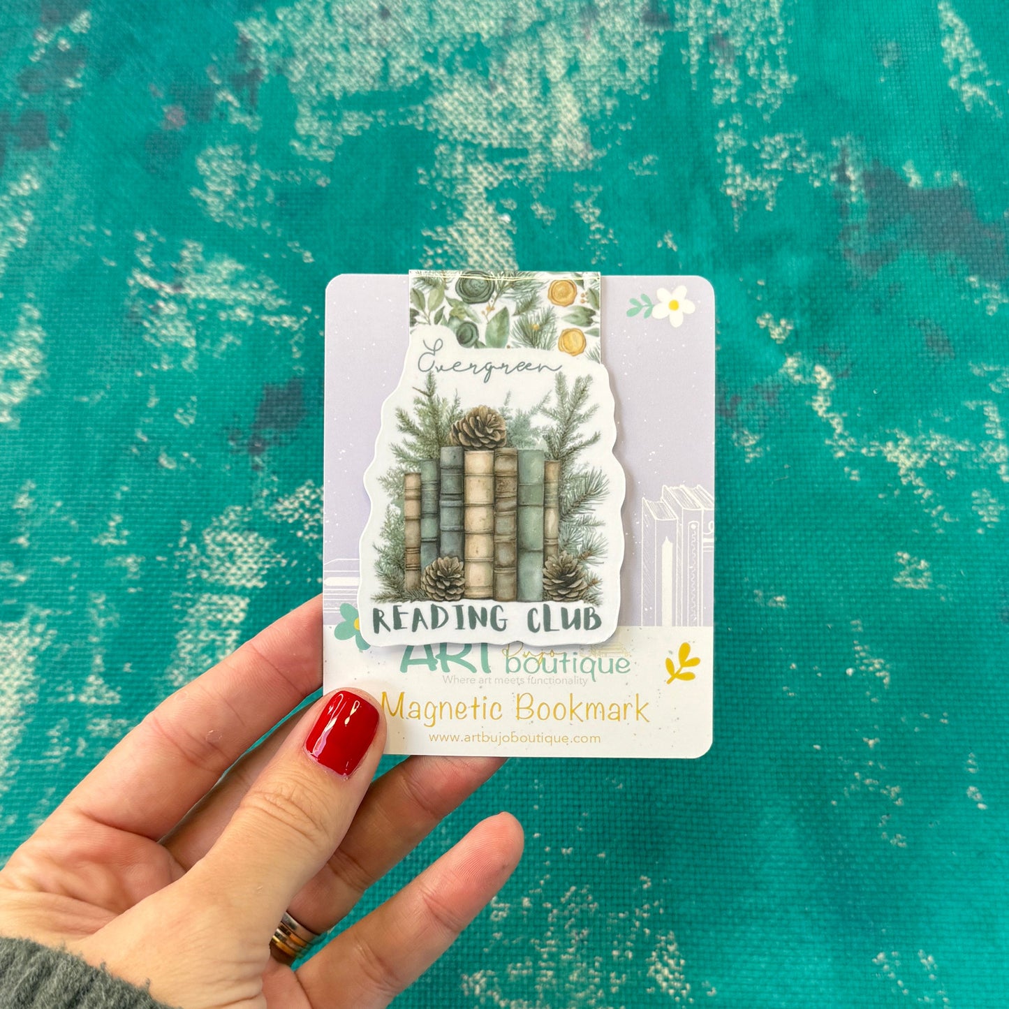 A hand with red nail polish holds a decorative magnetic bookmark card featuring stacked books, pine trees, and the text 'READING CLUB' and 'AKI Boutique'. The background is a textured teal-green surface.