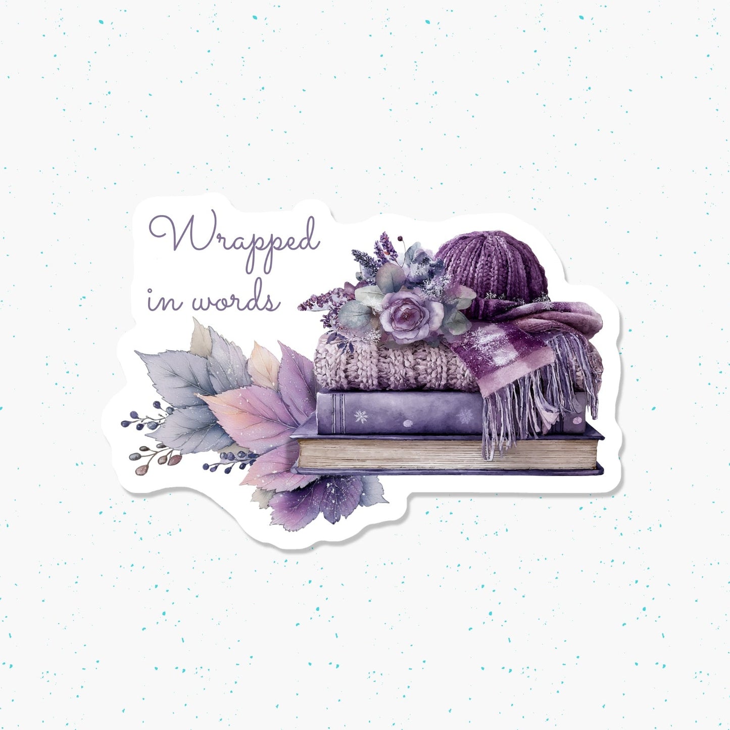 A die-cut sticker featuring a watercolor illustration of a purple knit hat, scarf, flowers, leaves, and a book with the text 'Wrapped in words' on a white speckled background.