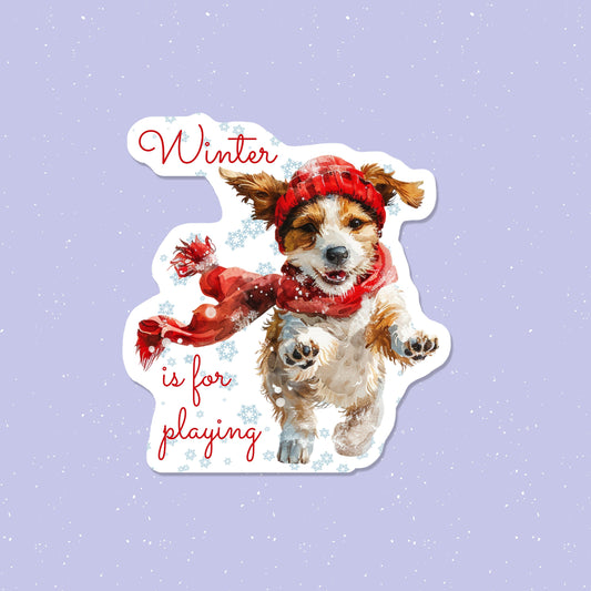 Sticker of a dog wearing a red hat and scarf with 'Winter is for playing' text on a light purple background.