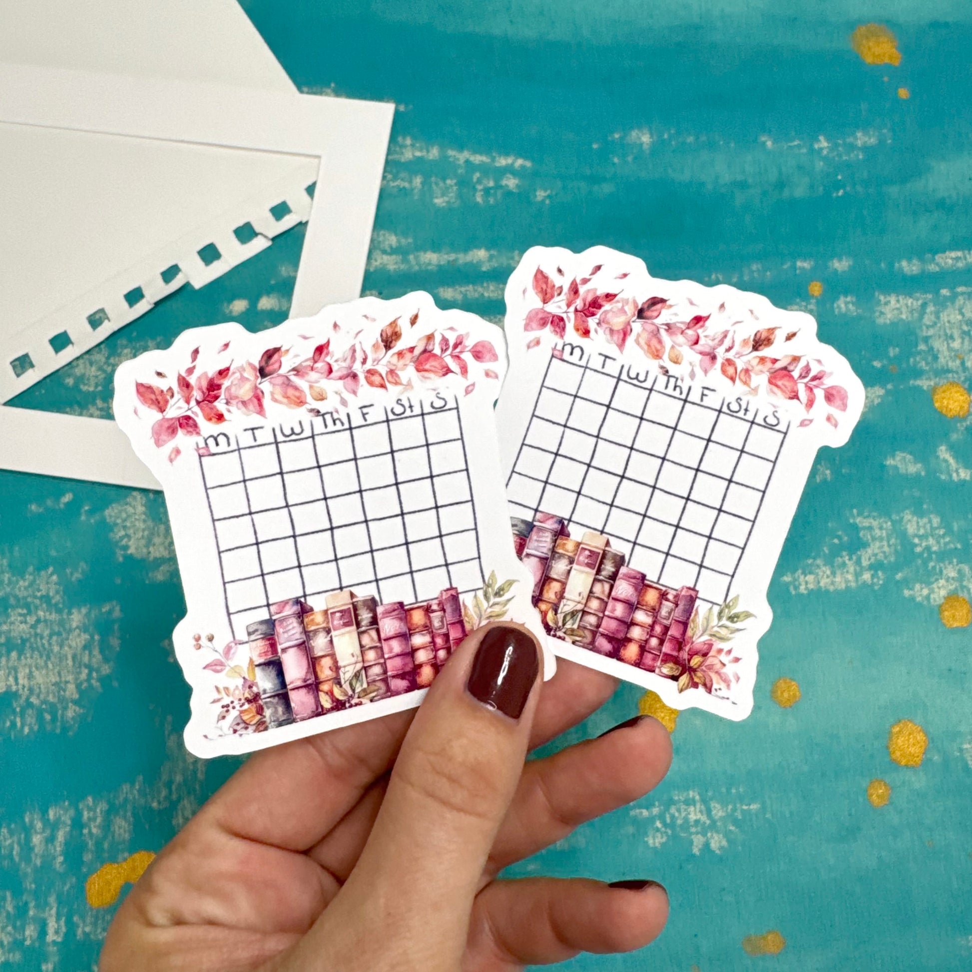 Hand holding two decorative planner stickers with floral and grid designs on a textured blue background.