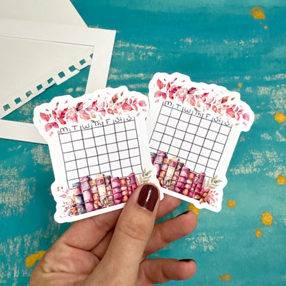 Hand holding two decorative planner stickers with floral and grid designs on a textured blue background.