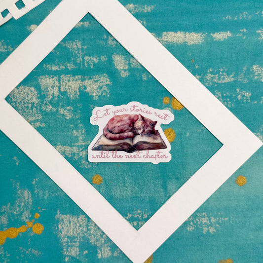 Sticker of a cat on an open book with text 'Let your stories rest until the next chapter' on a textured teal background.