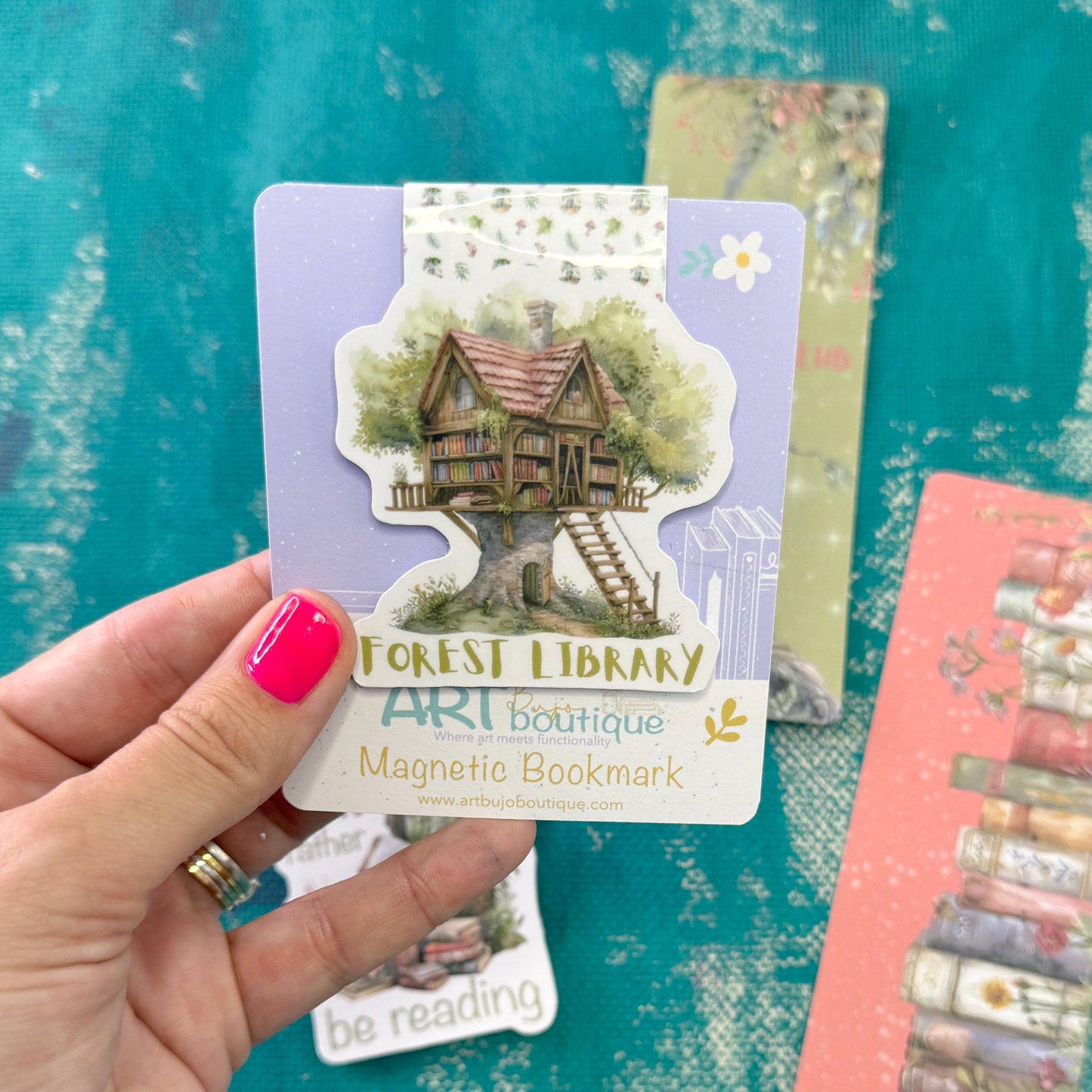 A hand with bright pink nail polish holds a die-cut magnetic bookmark featuring an illustration of a treehouse library. The bookmark card is lavender with gold and white text reading 'Forest Library Art Boutique Magnetic Bookmark.