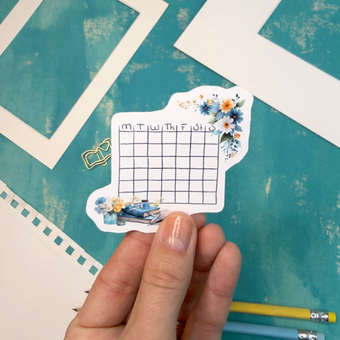 Small grid planner sticker with floral designs held by a hand on a textured blue surface.