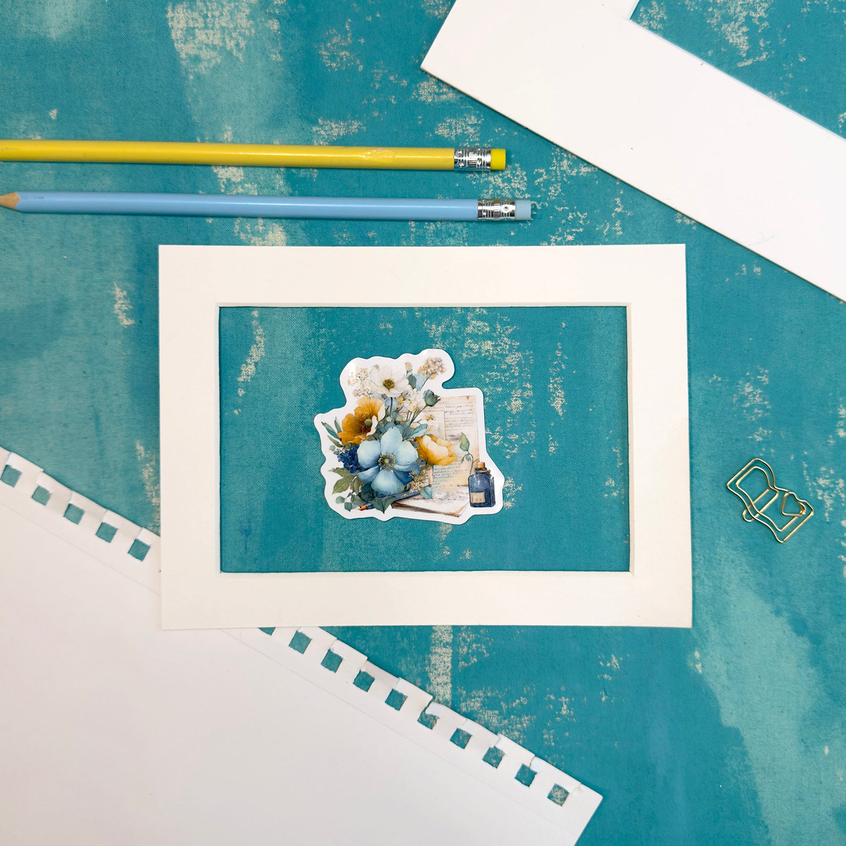 Framed artwork with a teddy bear and flowers on a textured blue surface with pencils.