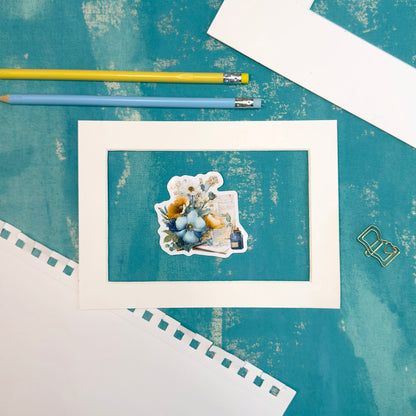 Framed artwork with a teddy bear and flowers on a textured blue surface with pencils.