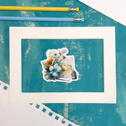 Framed artwork with a teddy bear and flowers on a textured blue surface with pencils.