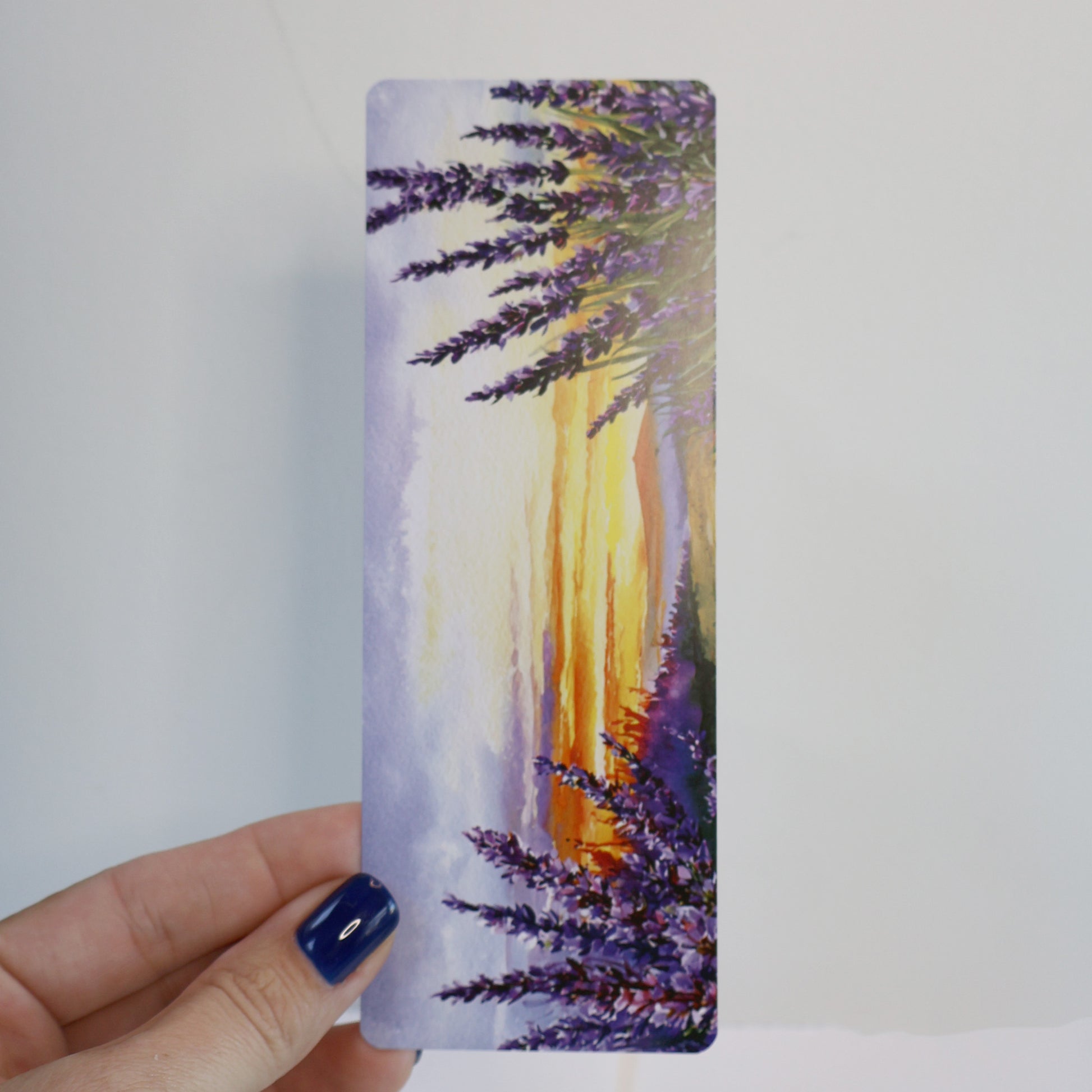 Hand holding a bookmark with a sunset and lavender design against a white background