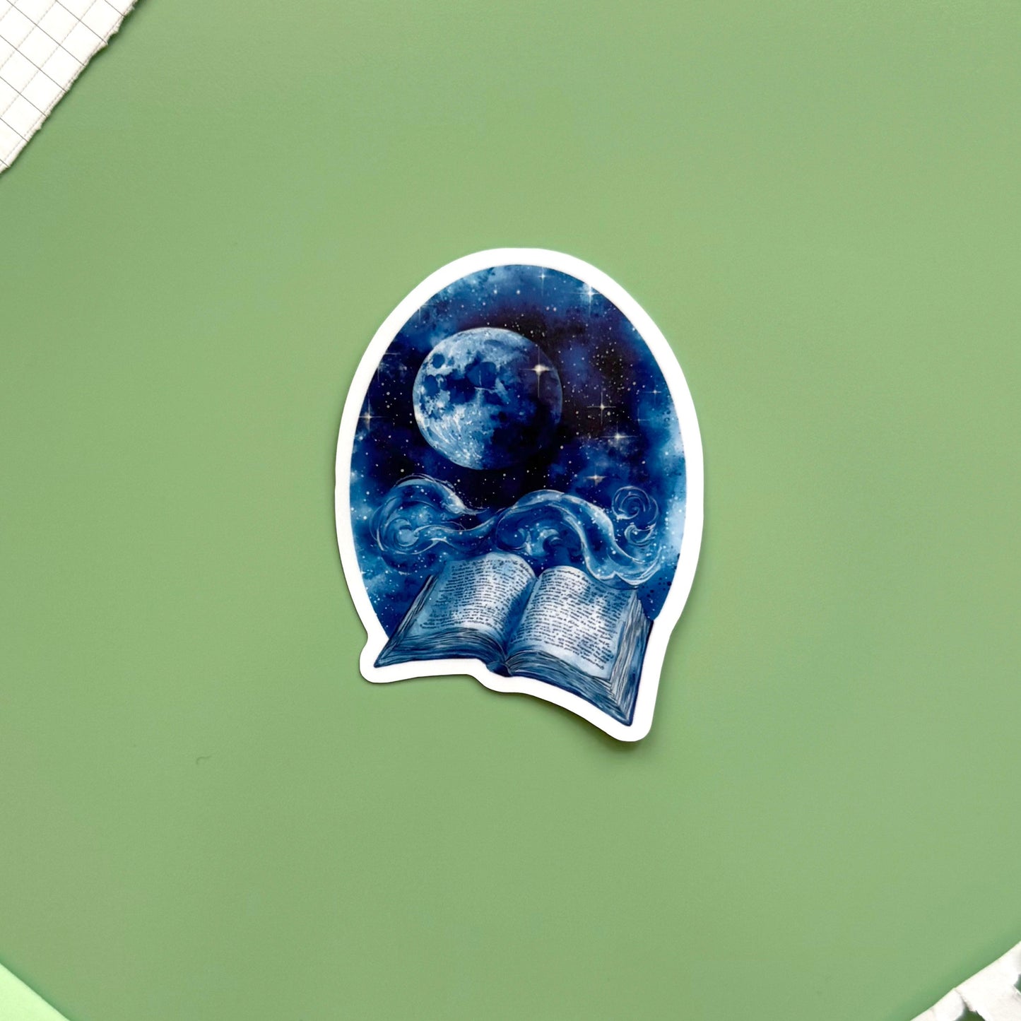 A die-cut sticker featuring a blue night sky illustration with a full moon and an open book, placed on a sage green background with notebook corners visible.