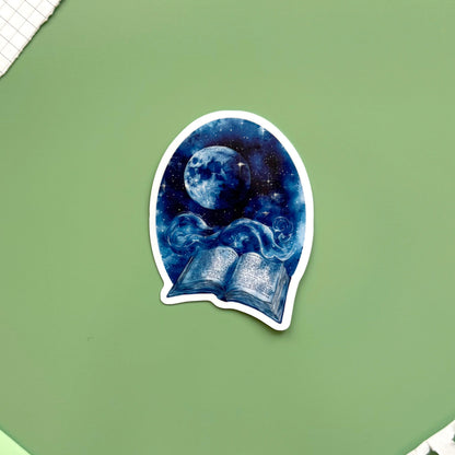 A die-cut sticker featuring a blue night sky illustration with a full moon and an open book, placed on a sage green background with notebook corners visible.