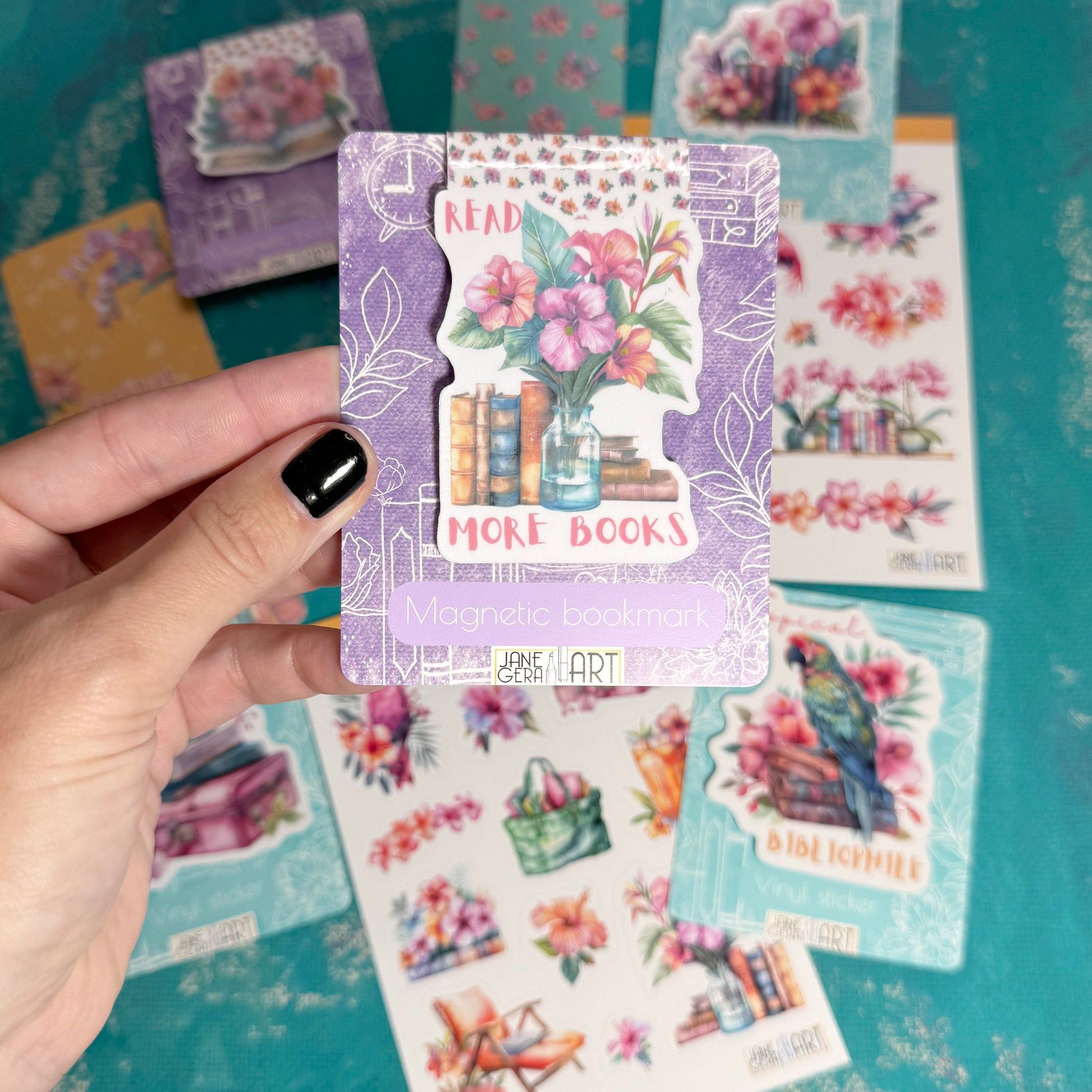 A hand with black nail polish holds a lavender decorative sticker card featuring a floral book illustration and the words 'READ MORE BOOKS'. Multiple other colorful sticker sheets and cards are scattered on a teal textured background.