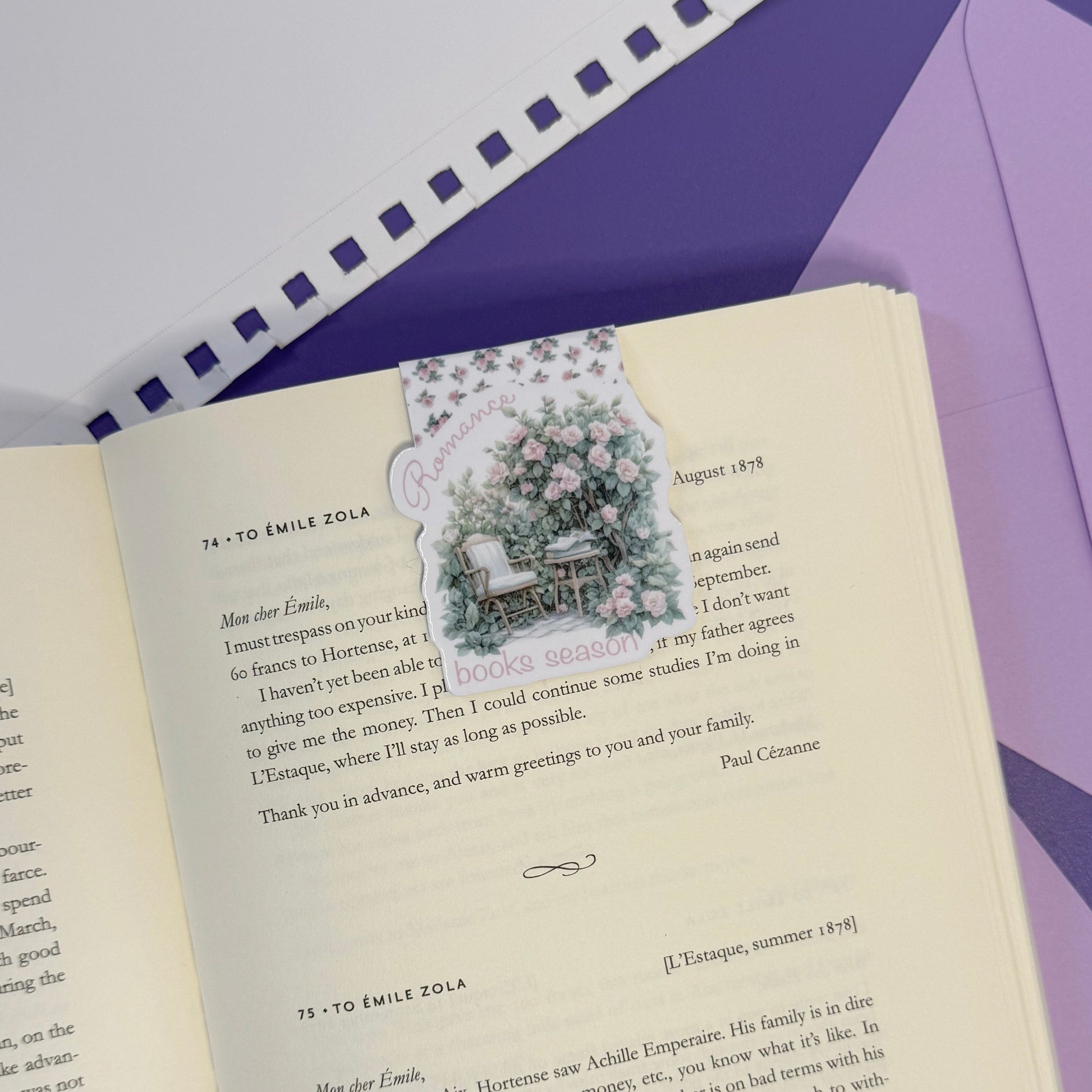 An open vintage-style book with printed text and a small illustrated postcard featuring a woman and children at a table, placed on a purple surface next to a matching envelope.