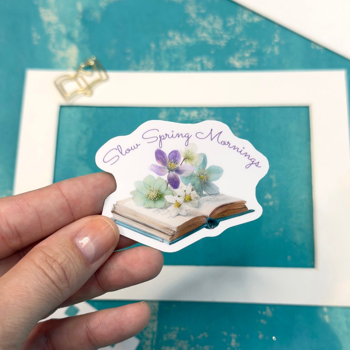 Hand holding a sticker with floral design and text 'Sweet Spring Mornings' against a teal background with a framed picture and pencil.