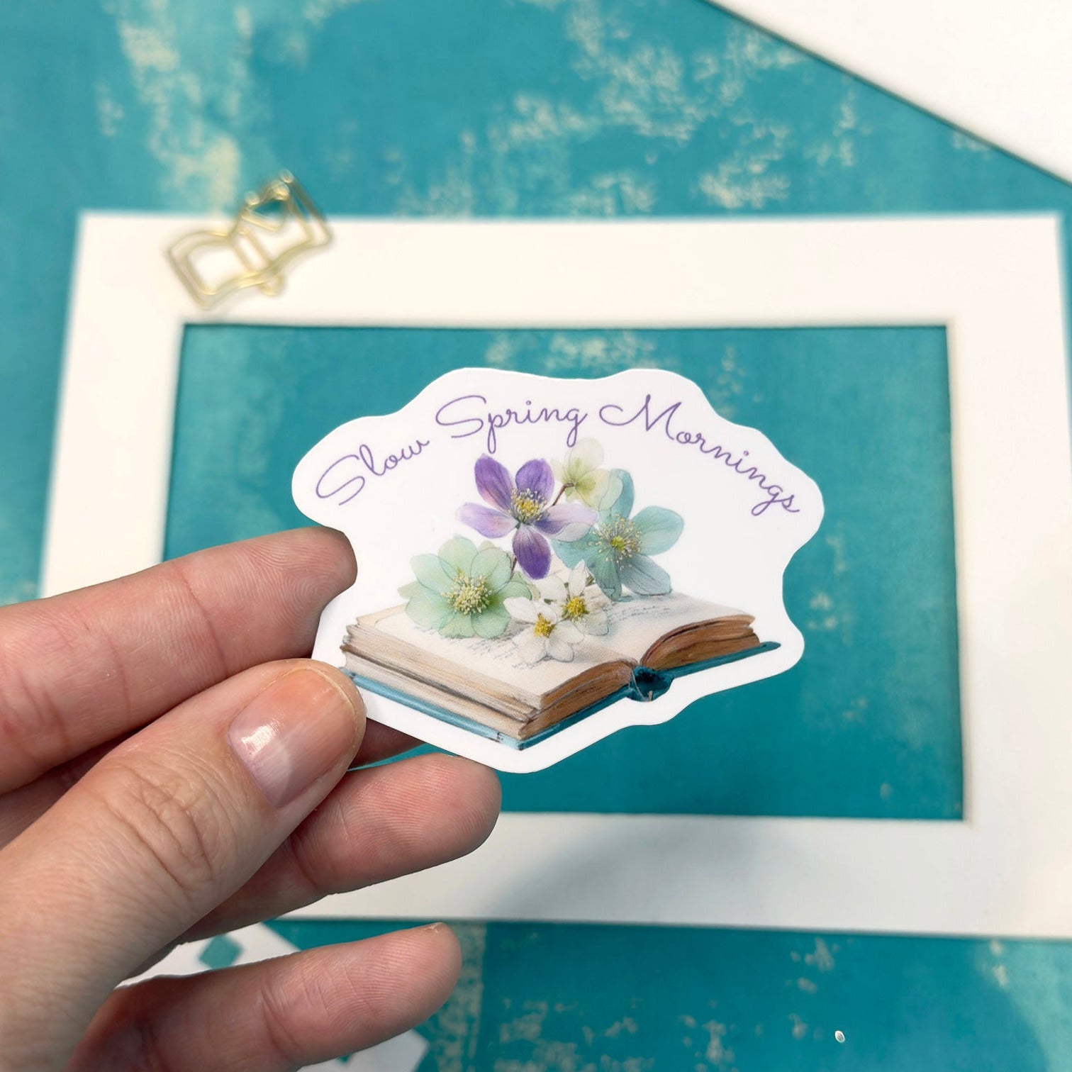 Hand holding a sticker with floral design and text 'Sweet Spring Mornings' against a teal background with a framed picture and pencil.