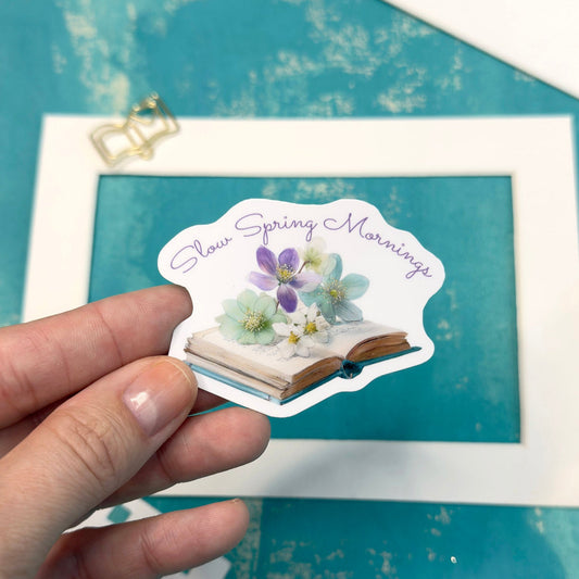 Hand holding a sticker with floral design and text 'Sweet Spring Mornings' against a teal background with a framed picture and pencil.
