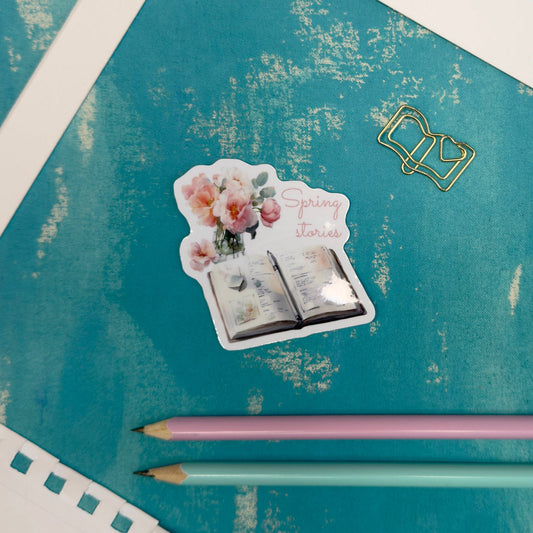 Sticker of a book with flowers and text on a textured blue surface with two pencils.