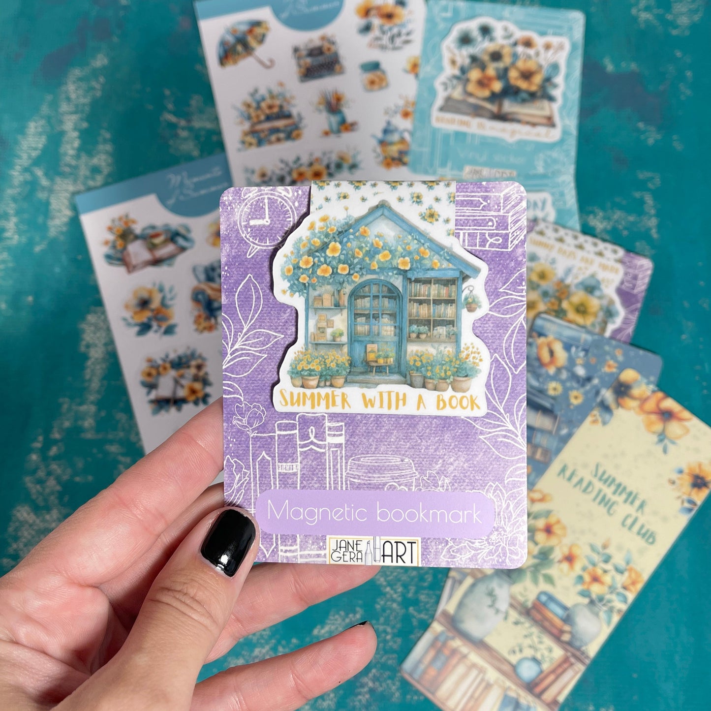 A hand with black nail polish holds a lavender 'Magnetic bookman' card featuring a whimsical house illustration, with several other decorative cards fanned out behind it on a textured teal background.
