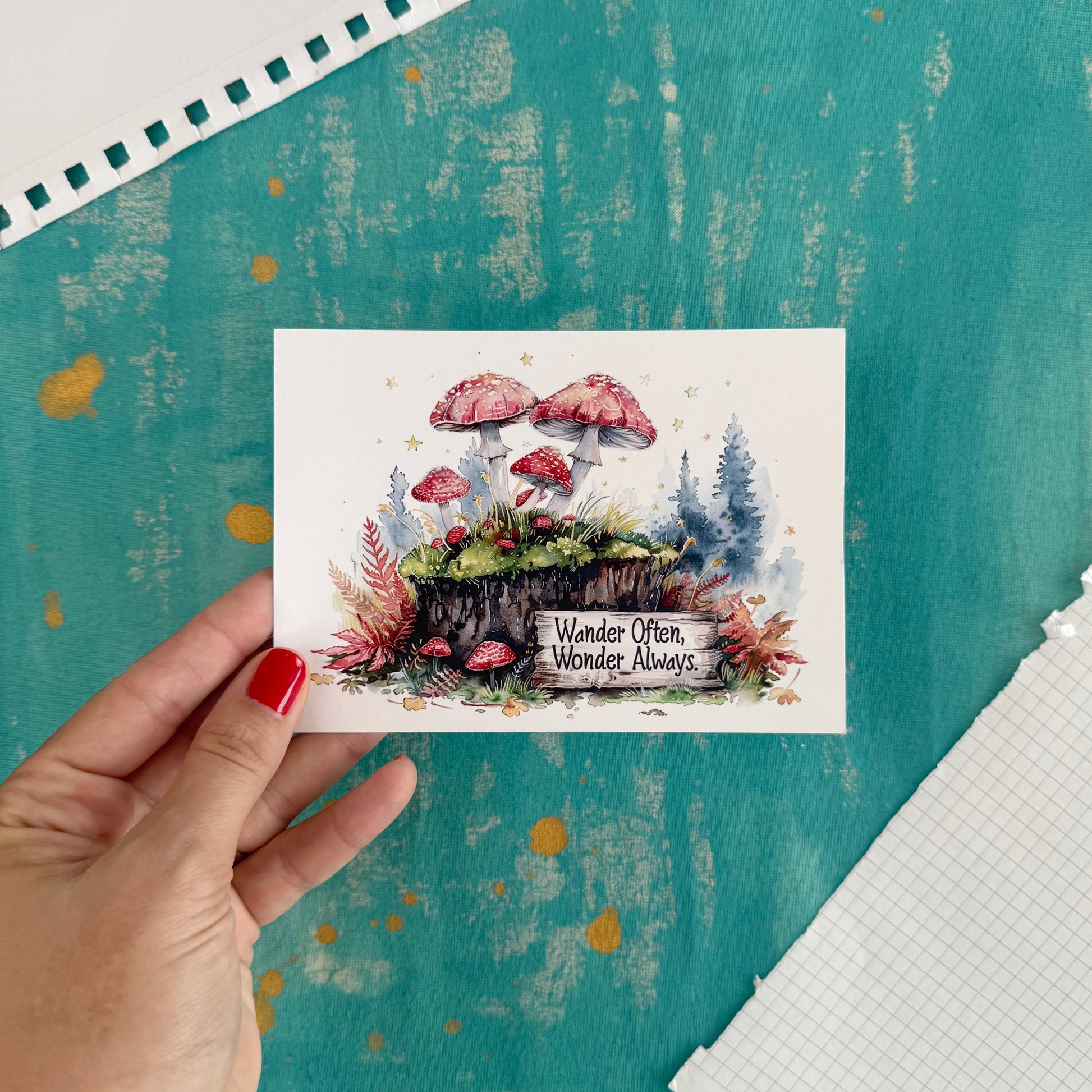 Hand holding a card with mushroom illustration and text 'Wander Often, Wonder Always' against a textured teal background.