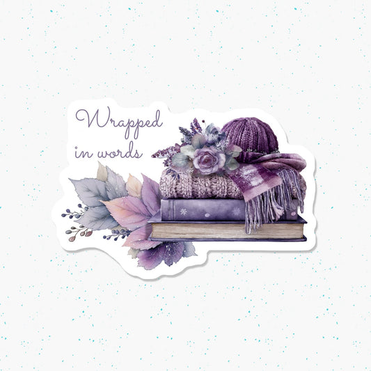 A die-cut sticker featuring a watercolor illustration of a purple knit hat, scarf, flowers, leaves, and a book with the text 'Wrapped in words' on a white speckled background.